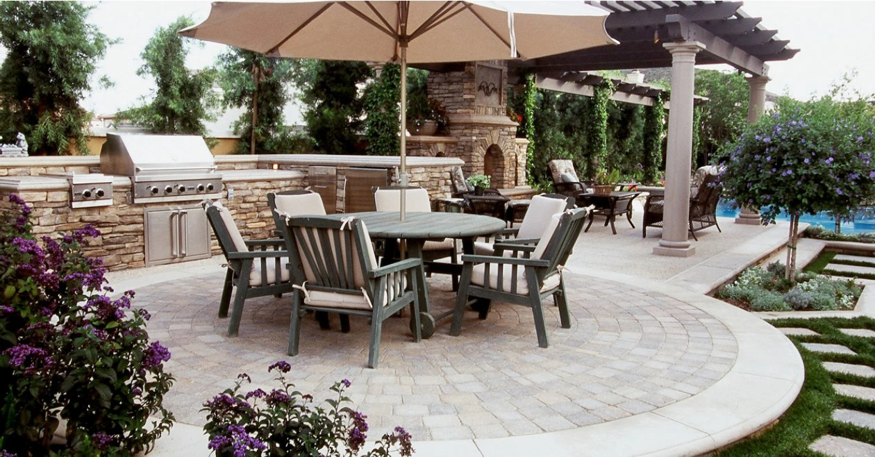 Custom concrete patio in Southlake TX with outdoor kitchen featuring stone veneer, curved paver design, pergola shade structure, and professional landscaping integrating purple flowering plants and modern outdoor furniture.